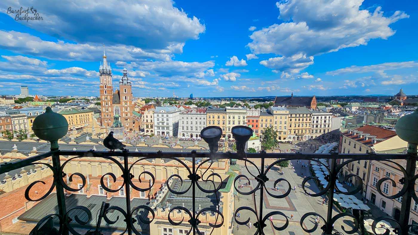 A panoramic view over Kraków's Main Market Square from high above the surrounding rooftops, framed in the foreground by an ornate black metal balcony railing. The twin unequal towers of St Mary's Basilica rise prominently on the left, overlooking rows of colourful historic façades and the busy square below. A vivid blue sky filled with large white clouds makes up almost half the scene, giving the sprawling city view a bright, expansive feel.