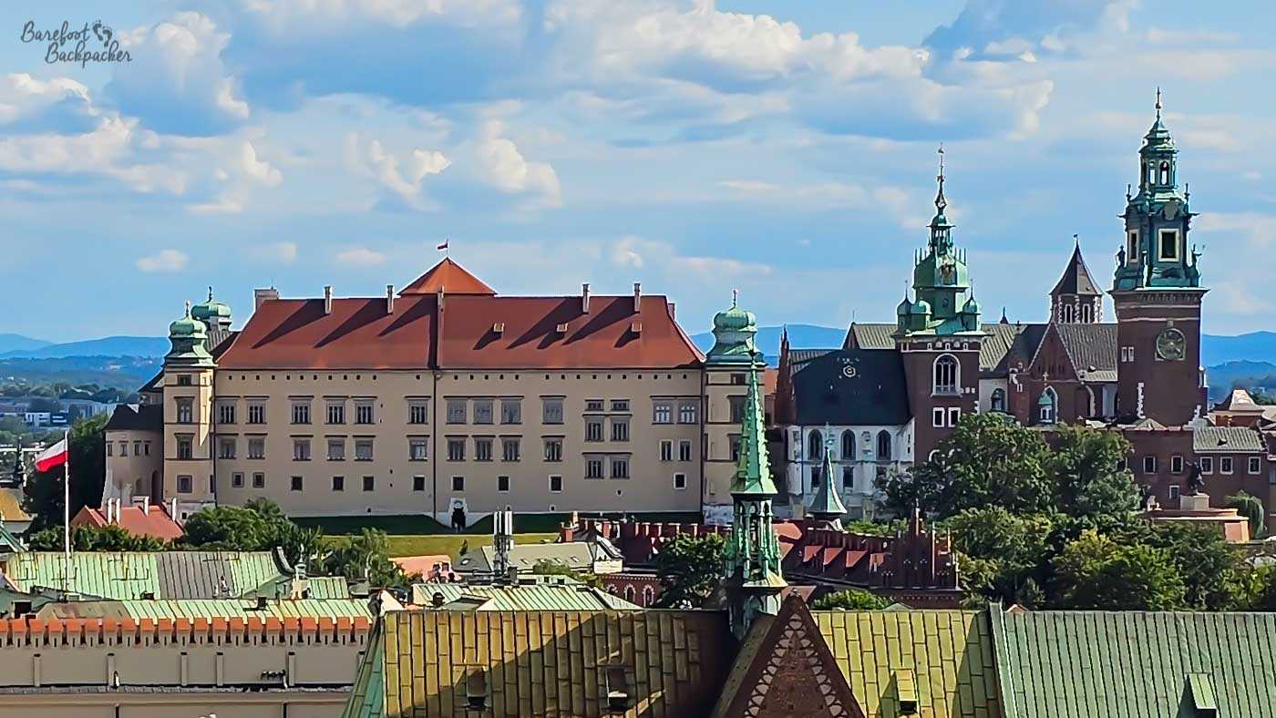 A view across the rooftops of Kraków towards Wawel Castle and Cathedral on a bright summer afternoon. The long pale façade and red roof of the castle dominate the left of the skyline, while the cathedral complex to the right is a jumble of red brick, dark roofs and tall towers topped with green-patinated copper. Colourful roofs and spires fill the foreground, with hazy hills visible beyond the city.