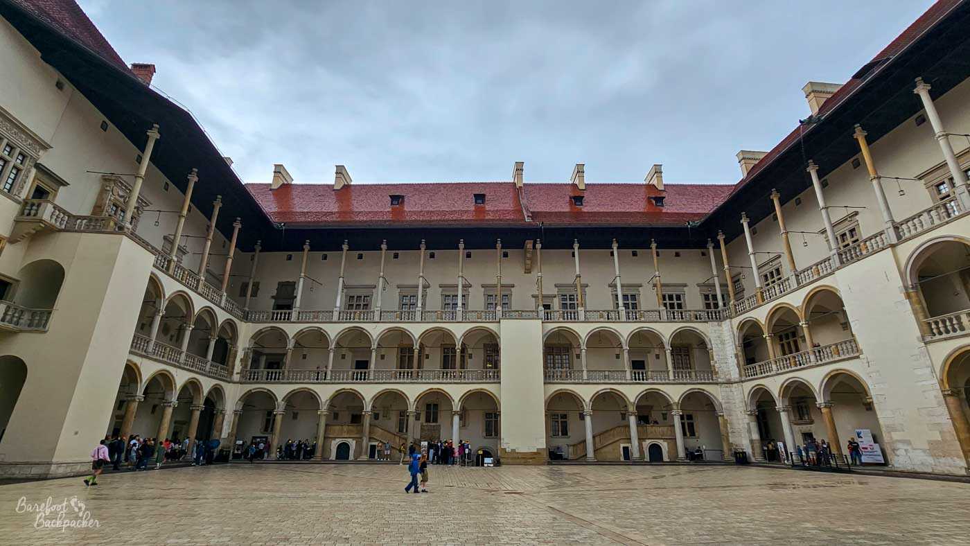 The large Renaissance courtyard of Wawel Castle in Kraków, enclosed on three sides by several storeys of pale arcaded galleries. Slender columns support repeated rounded arches around the courtyard, beneath a steep red-tiled roof dotted with chimneys and small windows. Visitors gather in small groups around the edges of the broad stone-paved open space beneath an overcast sky.