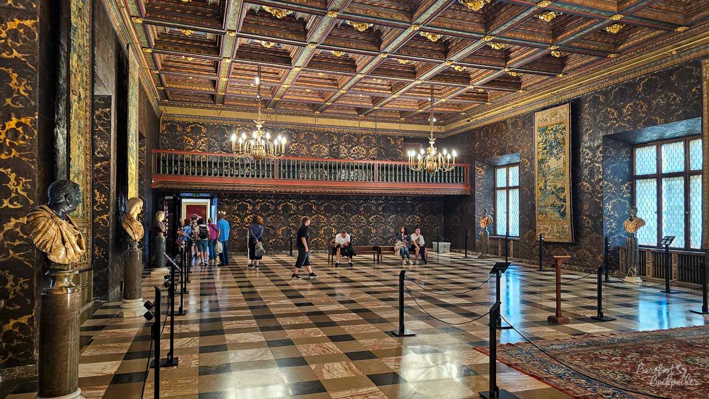 An elaborate historic interior in Kraków's Wawel Castle, with a large chequered stone floor surrounded by dark walls richly patterned with gold decoration. An intricately panelled and painted wooden ceiling stretches overhead, lit by chandeliers, while busts, a large tapestry and other exhibits stand around the room. Several visitors wander or sit on benches, their small scale emphasising the size and grandeur of the chamber.