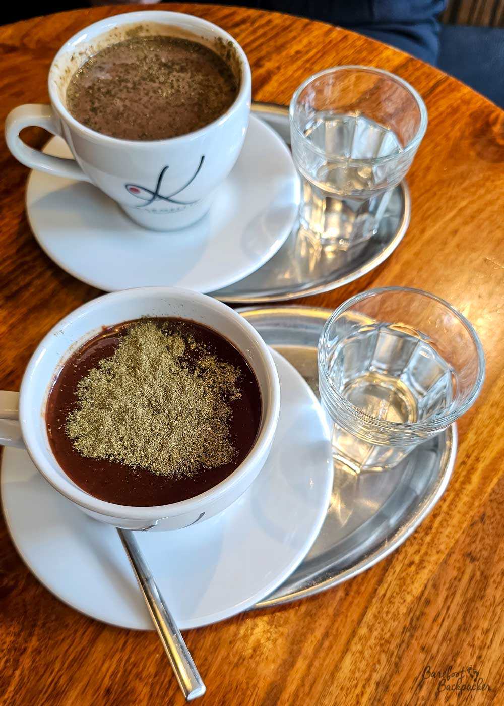 Two cups of thick hot chocolate sit on metal trays on a polished wooden café table, each accompanied by a small glass of water. The nearer drink is dark and glossy with a generous dusting of fine golden-coloured powder across its surface, while the second cup behind is lighter and more textured. The close-up view emphasises just how rich and substantial the drinks are.