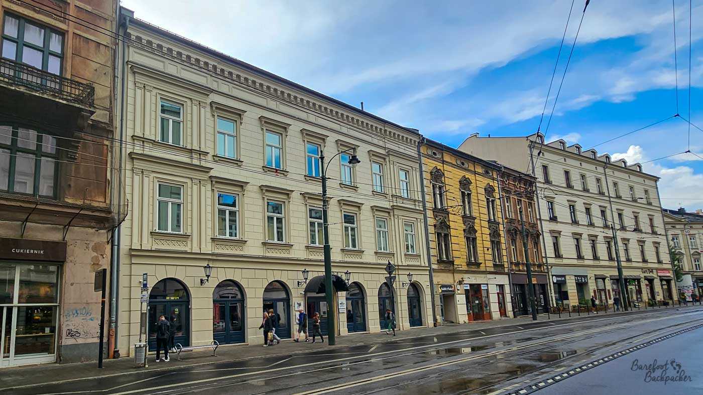 A row of ornate historic buildings lines a street in central Kraków, with cream, yellow and weathered stone façades rising above shops at ground level. Tram tracks run along the wet road in front, while a network of tram wires crosses the blue-and-cloudy sky overhead. A handful of pedestrians walk along the pavement, giving the scene the feel of an ordinary city street surrounded by grand architecture.