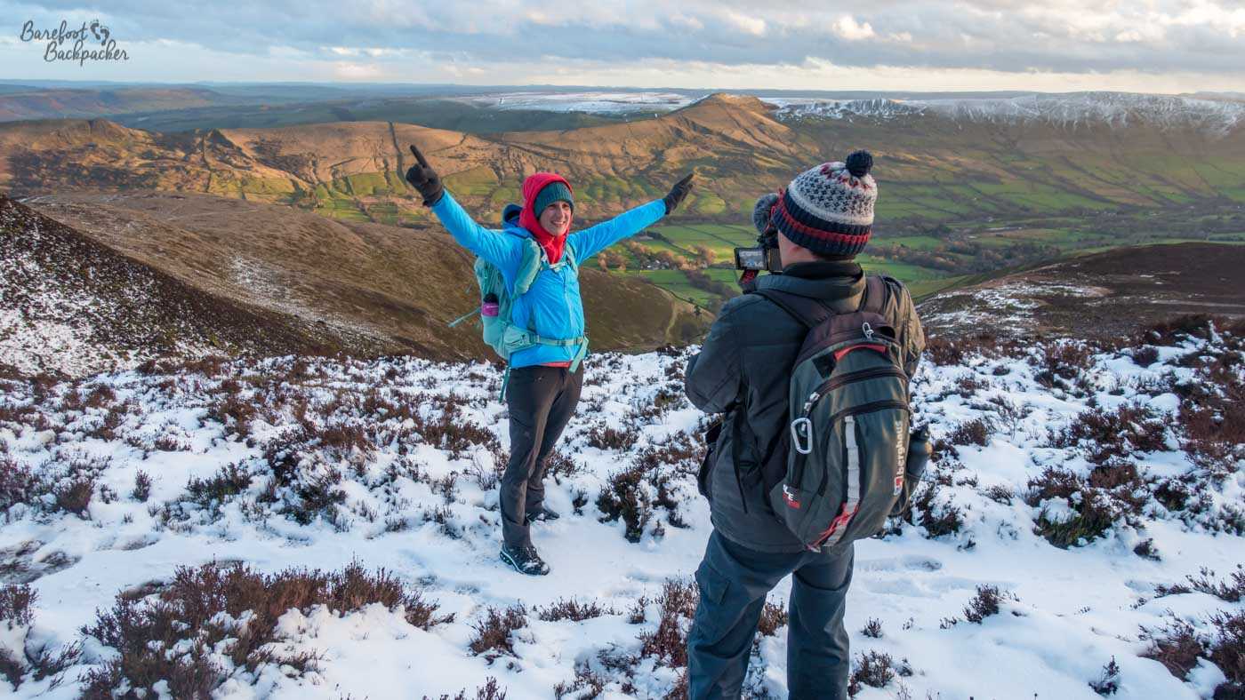 A hiker in a bright blue jacket and red hat stands with both arms raised on a snow-dusted moorland hillside, while another hiker wearing a large backpack photographs them. Behind them, the ground drops into a broad green valley surrounded by rolling brown hills, with streaks of snow along the higher ridges beneath a cloudy winter sky.