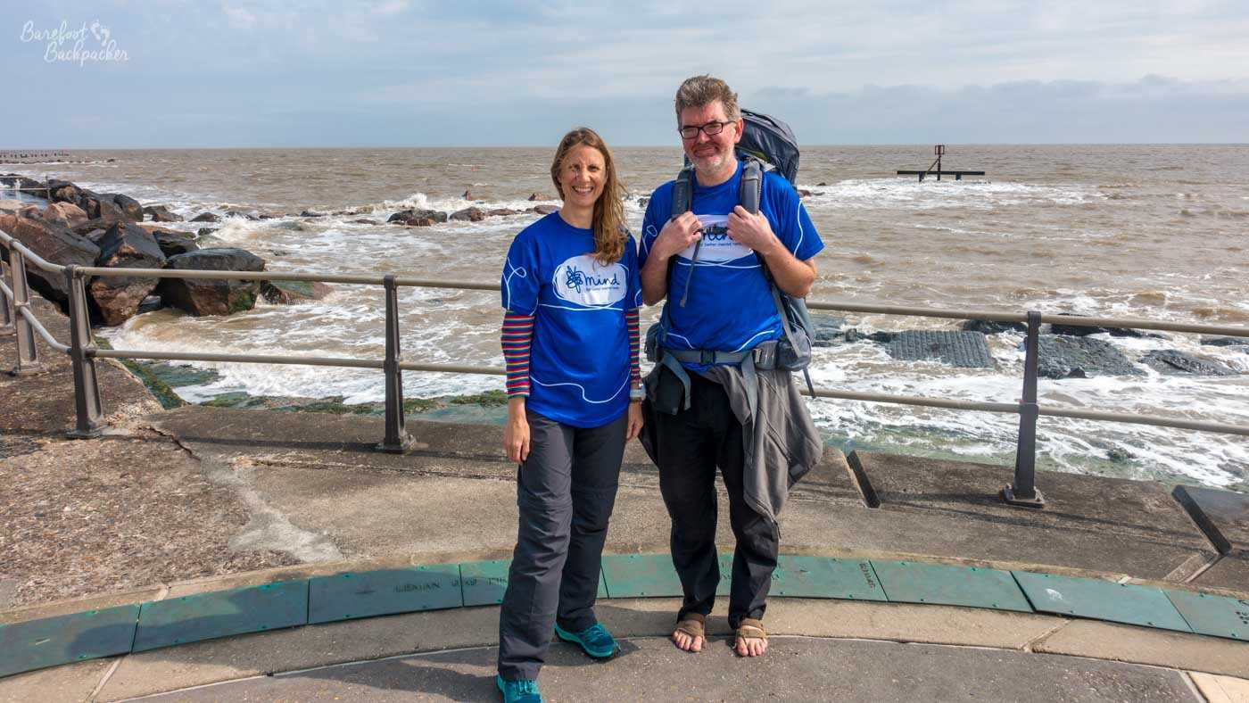 A man and a woman, both wearing blue t-shirts and dark trousers, stand on a paved walkway by the sea. The woman is on the left, with long brown hair and a striped long-sleeved shirt visible under her t-shirt. The man is on the right, wearing glasses and a backpack, and has a sweater tied around his waist. He is also wearing crocheted sandals, while the woman wears athletic shoes. They are both smiling at the camera. The sea behind them is choppy, with waves breaking against a rocky shoreline and a metal railing. The sky is overcast. A marker buoy is visible in the distance.
