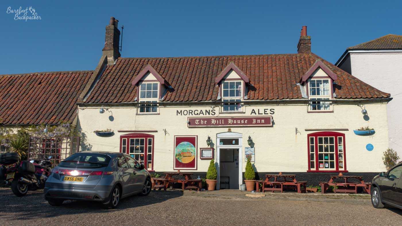 The front of the Hill House Inn in Happisburgh on a clear sunny day. The traditional cream-painted pub has a red tiled roof, dormer windows and “Morgans Ales” painted across its frontage, with wooden picnic tables and several parked vehicles outside beneath a deep blue sky.