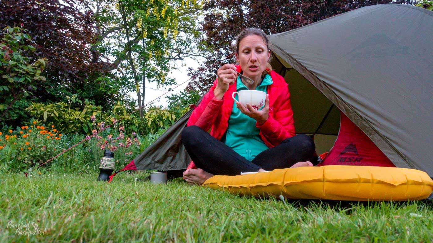 A hiker sits cross-legged and barefoot on the grass outside a small pitched tent, eating from a white mug with a spoon. They wear black leggings, a turquoise top and bright red jacket, with a yellow sleeping mat in the foreground and flowers, shrubs and trees surrounding the campsite.