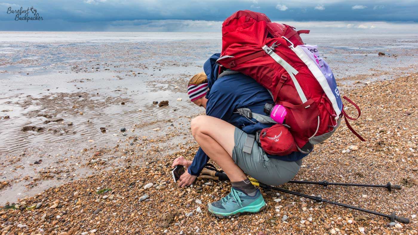 A hiker crouches on a pebbly beach beneath an enormous red backpack, examining something among the stones near the water’s edge. Walking poles lie on the beach beside them, while shallow tidal water stretches towards the horizon under a dark, heavily clouded sky.