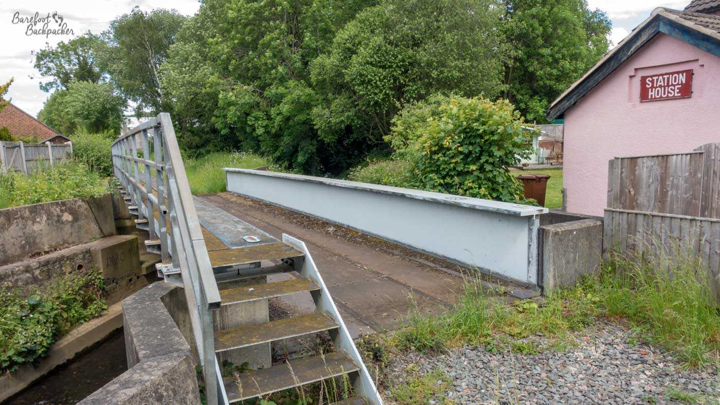 The remains of the former Little Eaton railway station, with an old platform beside a narrow water channel and metal steps leading down in the foreground. A small pink building marked “Station House” stands to the right, while trees and overgrown vegetation surround the disused railway site.