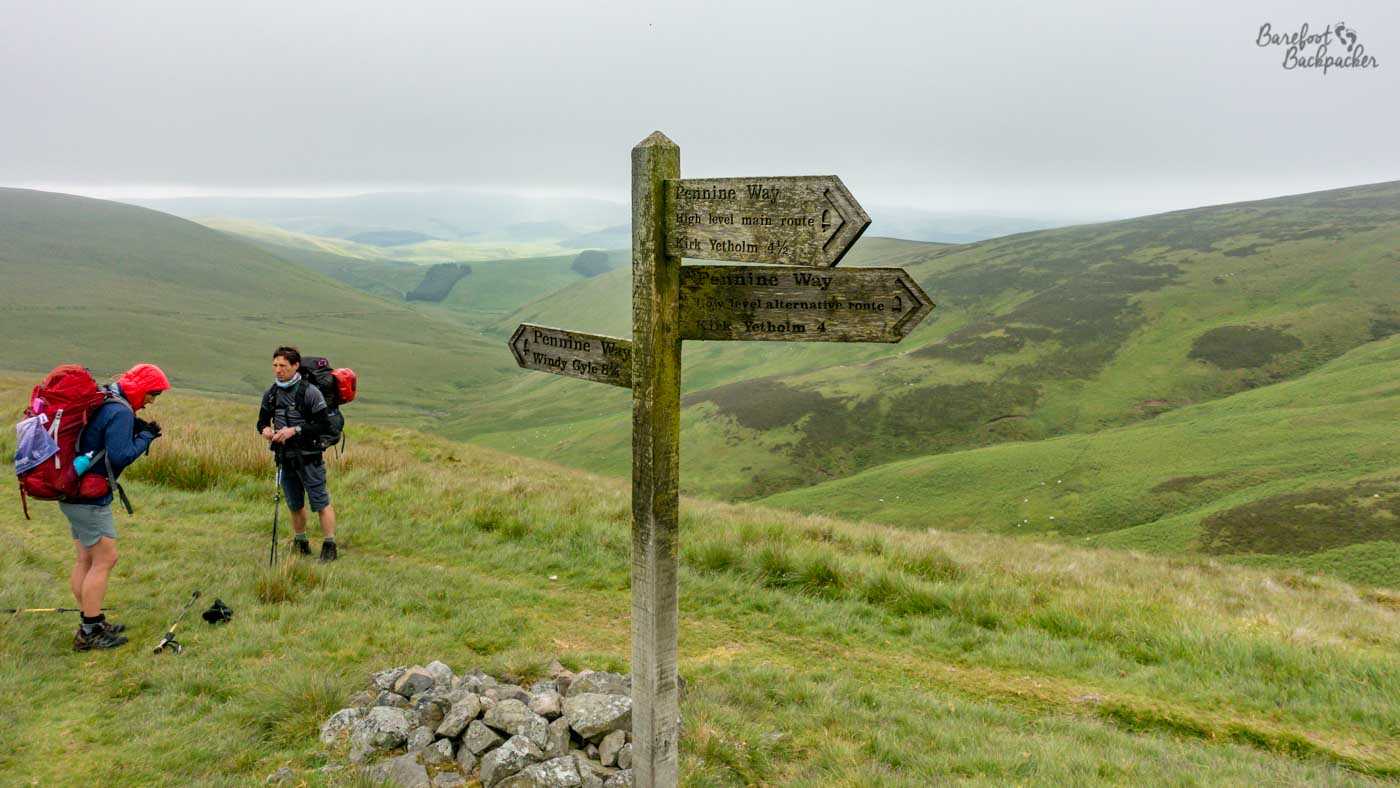 A weathered wooden Pennine Way signpost stands high in the green, treeless hills near the Scottish border, pointing towards Kirk Yetholm and back towards Windy Gyle. Two backpackers have stopped beside the path to the left, dwarfed by the broad rolling landscape disappearing into low grey cloud.