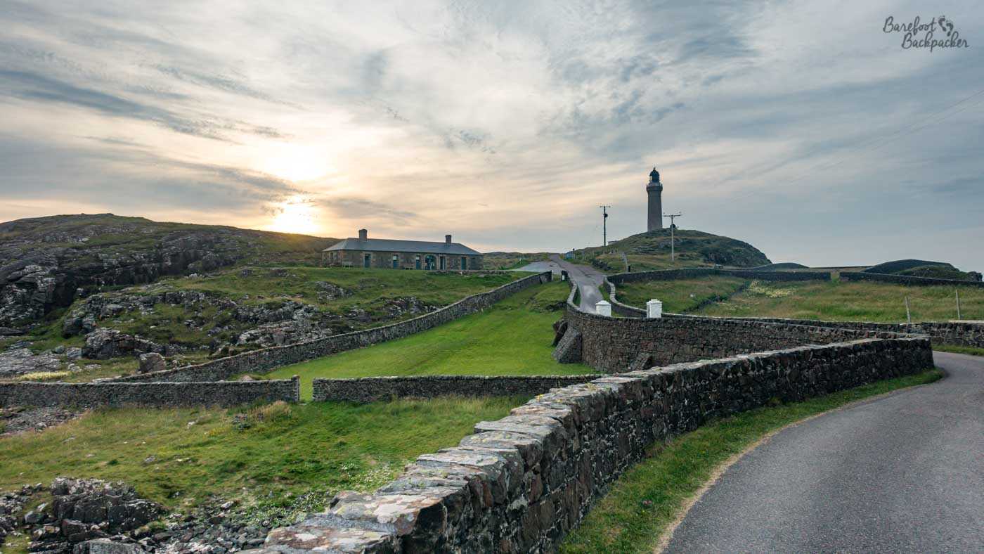 Ardnamurchan Lighthouse stands on a rocky headland beneath a softly clouded evening sky, reached by a narrow road winding between low stone walls. The setting sun glows behind the rugged landscape to the left, with a long low building below the lighthouse and the sea just visible beyond the headland.