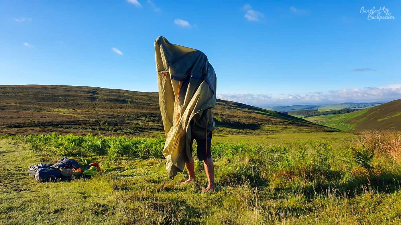 A barefoot person stands on a grassy hillside almost completely enveloped by a large collapsed green tent, with only their lower legs and feet emerging beneath it. Camping equipment lies in a heap nearby, while rolling green and brown hills stretch into the distance under a vivid blue sky.